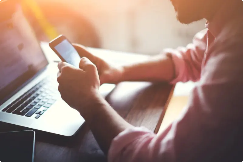 Handen met een telefoon en een laptop op de achtergrond, passend bij de voordelen van periodiek beleggen